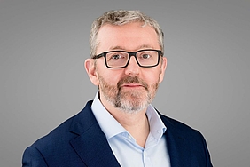 Älterer Mann mit Brille, grauen Haaren und Bart mit weißem Hemd und dunklem Sakko